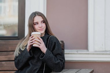 Bir ceket genç güzel bin yıllık kız kahve bir kağıt fincan ile bir outdor bankta oturuyor. Sonbahar veya ilkbahar günü, serin hava.