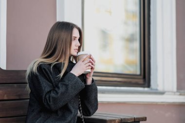 Bir ceket genç güzel bin yıllık kız kahve bir kağıt fincan ile bir outdor bankta oturuyor. Sonbahar veya ilkbahar günü, serin hava.