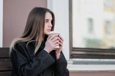 Bir ceket genç güzel bin yıllık kız kahve bir kağıt fincan ile bir outdor bankta oturuyor. Sonbahar veya ilkbahar günü, serin hava.