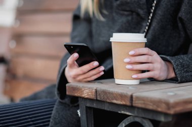 Açık bir kafebankında kahverengi bir kağıt fincan kahve ile kadın ellerini yakın çekim. Akıllı telefon ve Internet kullanır.
