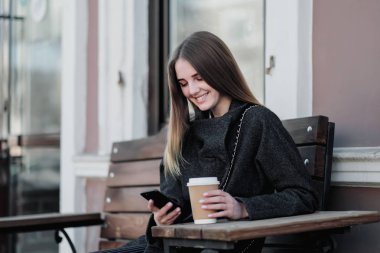 Bir ceket genç güzel bin yıllık kız kahve bir kağıt fincan ile bir outdor bankta oturuyor. Sonbahar veya ilkbahar günü, serin hava.