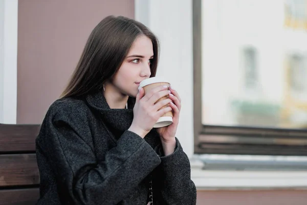 Mladá krásná tisíciletí v kabátě sedí na lavici s papírovým šálkem s kávou. Podzimní nebo jarní den, chladné počasí. — Stock fotografie