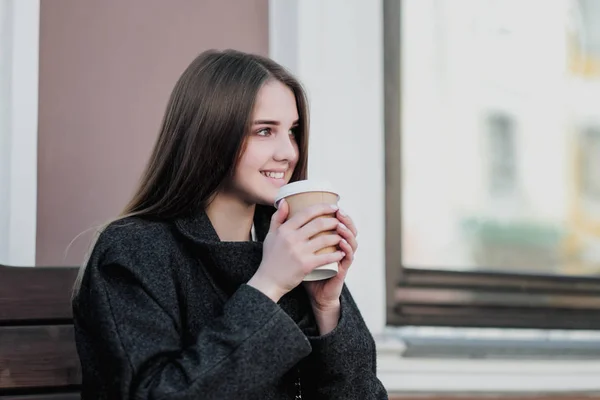 Mladá krásná tisíciletí v kabátě sedí na lavici s papírovým šálkem s kávou. Podzimní nebo jarní den, chladné počasí. — Stock fotografie
