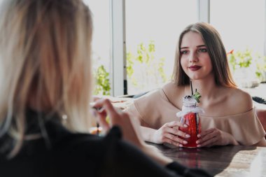 Elbiseler içinde iki genç akıllı güzel kız büyük bir pencerenin karşısında bir restoran masada oturup. Tübüllü bardaklardan kırmızı ve mavi kokteyller için.