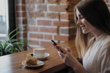 Bir çatı tarzı kafede genç bir güzel esmer kız pencerenin yanında bir masada oturur, kahve içecekler ve akıllı telefon kullanır, aramalar, fotoğraf çeker.