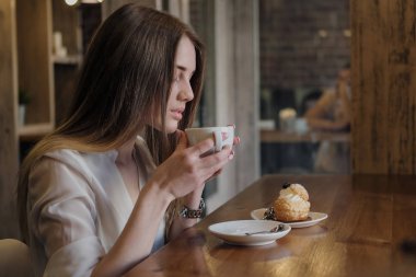 Bir çatı tarzı kafede Genç güzel esmer kız pencere nin yanında bir masada oturur, kahve içecekler ve tatlı kek yiyor.