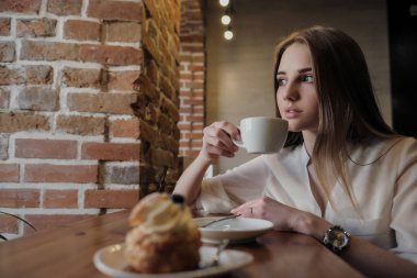 Bir çatı tarzı kafede Genç güzel esmer kız pencere nin yanında bir masada oturur, kahve içecekler ve tatlı kek yiyor.