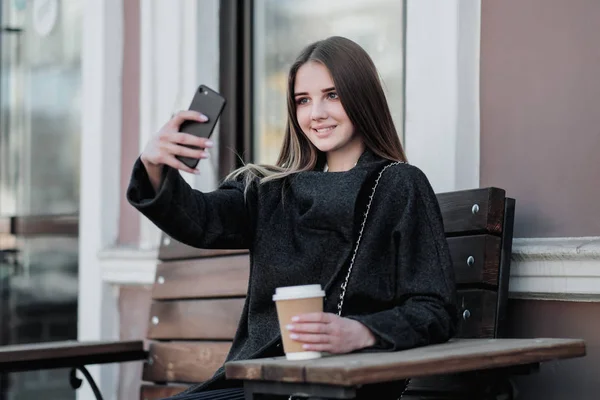 Jonge mooie Millenial meisje in een jas zit op een buitendor Bank met een papieren kopje koffie. Herfst-of lente dag, koel weer. — Stockfoto