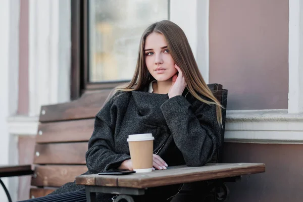 Das junge wunderschöne Millenium-Mädchen im Mantel sitzt mit einer Pappbecher Kaffee auf einer Bank im Außenbereich. Herbst- oder Frühlingstag, kühles Wetter. — Stockfoto