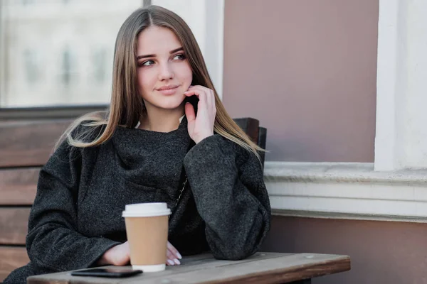 Jonge mooie Millenial meisje in een jas zit op een buitendor Bank met een papieren kopje koffie. Herfst-of lente dag, koel weer. — Stockfoto