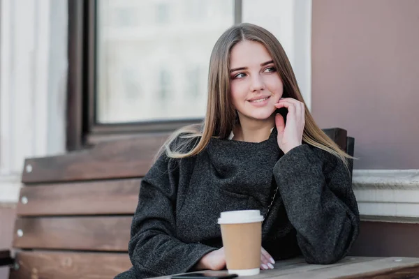 Jonge mooie Millenial meisje in een jas zit op een buitendor Bank met een papieren kopje koffie. Herfst-of lente dag, koel weer. — Stockfoto