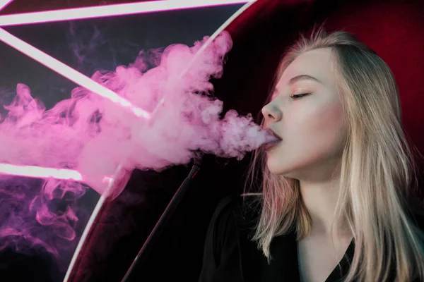 Schöne junge Frau Blondine atmet Rauch aus einer Wasserpfeife in einem Nachtclub vor dem Hintergrund von neonvioletten Lichtern. — Stockfoto