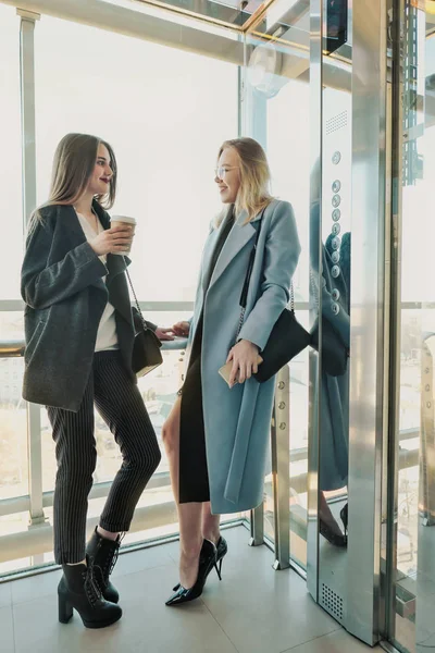 Two beautiful girls, a blonde and a brunette, are wearing a coat in a glass elevator with a window. Shopping center or office in the city. — Stock Photo, Image