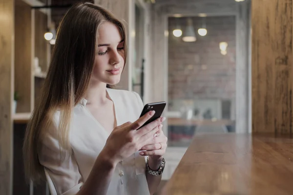 Bir çatı tarzı kafede genç bir güzel esmer kız pencerenin yanında bir masada oturur, kahve içecekler ve akıllı telefon kullanır, aramalar, fotoğraf çeker.
