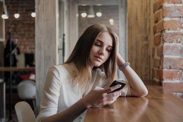 Młoda piękna brunetka dziewczyna w kawiarni w stylu loftu siedzi przy stole przy oknie, pije kawę i używa smartfona, połączeń, robi zdjęcia. — Zdjęcie stockowe