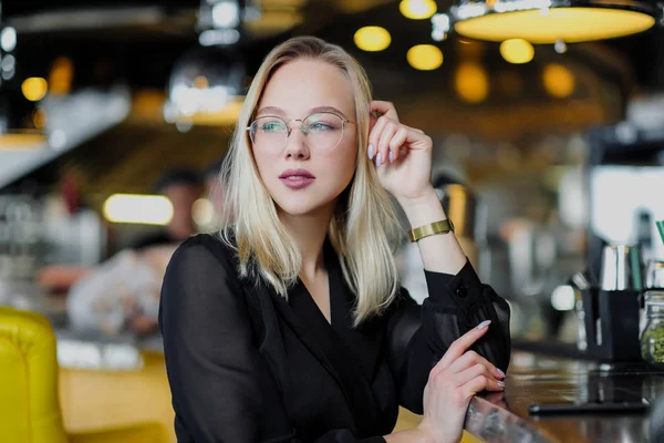 Uma jovem mulher loira bonita com óculos senta-se no bar em um interior amarelo. Bebe coquetel alcoólico com palha e hortelã verde . — Fotografia de Stock