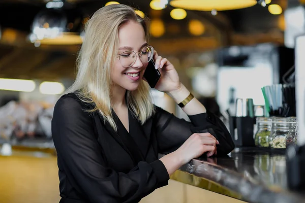 Una giovane bella donna bionda con gli occhiali si siede al bar in un interno giallo. Bevande cocktail alcolico con cannuccia e menta verde . — Foto Stock