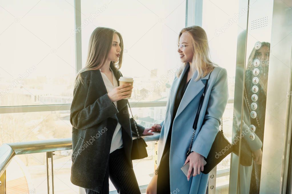 Two beautiful girls, a blonde and a brunette, are wearing a coat in a glass elevator with a window. Shopping center or office in the city.