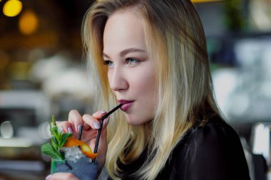 Gözlüklü genç ve güzel sarışın bir kadın barda sarı bir iç mekanda oturuyor. Bir saman ve yeşil nane ile Içecekler alkollü kokteyl.