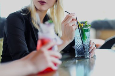 İki genç güzel kız, sarışın ve esmer, bir kafe veya restoranda bir masada oturup. İletişim ve konuşma, mavi ve kırmızı kokteyller içecekler içmek.