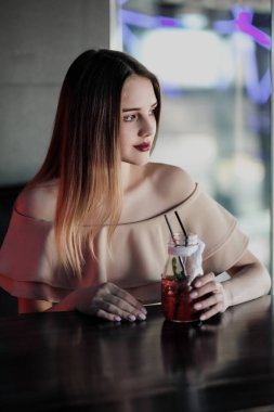 Neon ışıkları ile bir gece kulübünde genç güzel kahverengi saçlı kız cam oturur ve kırmızı bir kokteyl içecekler.