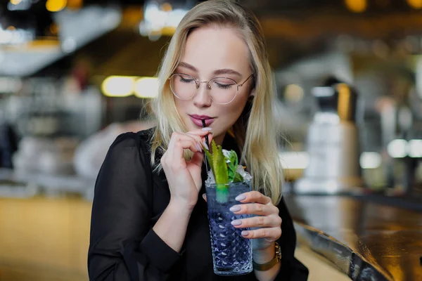 Una giovane bella donna bionda con gli occhiali si siede al bar in un interno giallo. Bevande cocktail alcolico con cannuccia e menta verde . — Foto Stock