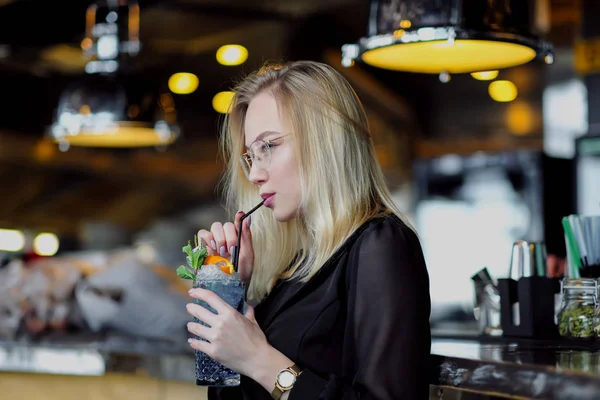 A young beautiful blond woman with glasses sits at the bar in a yellow interior. Drinks alcoholic cocktail with a straw and green mint.