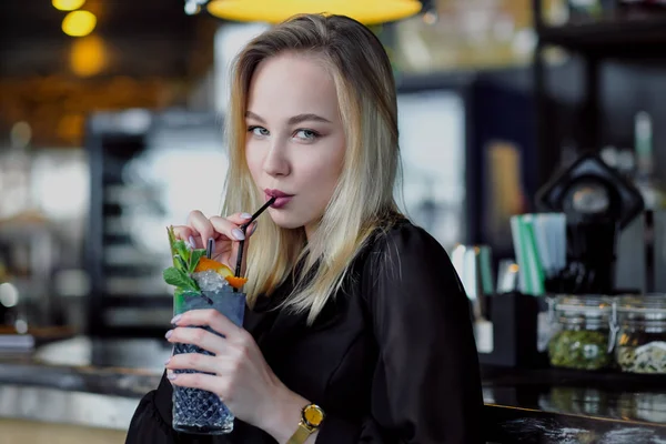 Una joven y hermosa rubia con gafas se sienta en el bar en un interior amarillo. Bebidas cóctel alcohólico con una paja y menta verde . —  Fotos de Stock