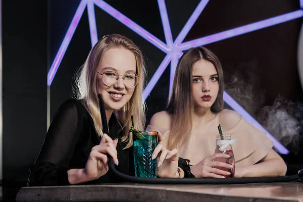 Two young smart beautiful girls in dresses sit at a table in a nightclub or hookah. They smoke hookah and smoke and drink cocktails from glasses with tubules, talk and laugh. — Stock Photo, Image