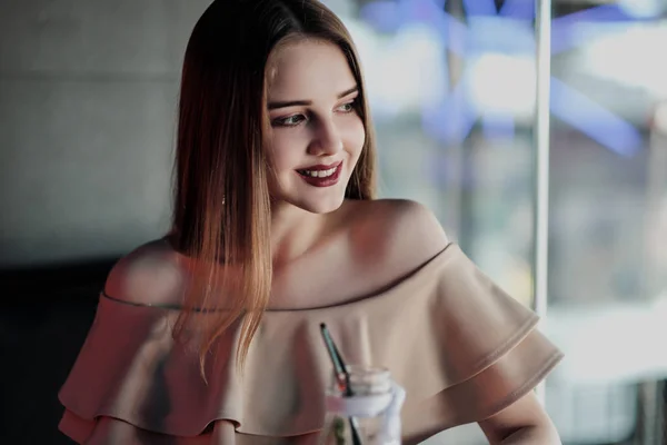 Giovane bella ragazza dai capelli castani in un nightclub con luci al neon si siede vicino al bicchiere e beve un cocktail rosso . — Foto Stock