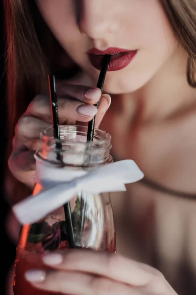 Young beautiful brown-haired girl in a nightclub with neon lights sits by the glass and drinks a red cocktail. — Stock Photo, Image