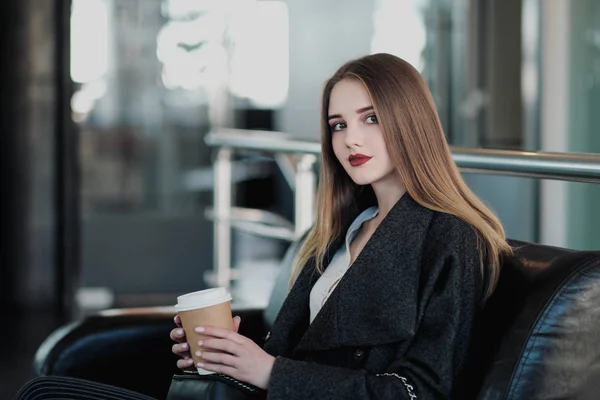 Młoda dziewczyna piękna brunetka w szarym płaszczu siedzi na ławce czeka. Picie kawy i trzymanie filiżanki papieru przy drinku. — Zdjęcie stockowe