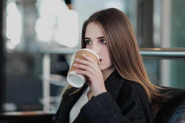 Mladá krásná brunetka v šedém kabátě sedí na lavičce a čeká. Popíjení kávy a držení papírového šálku s nápojem. — Stock fotografie