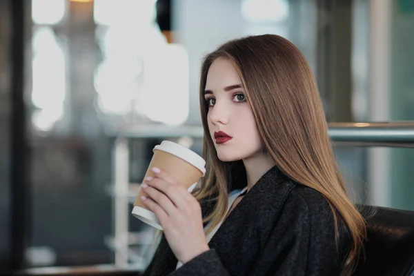 Mladá krásná brunetka v šedém kabátě sedí na lavičce a čeká. Popíjení kávy a držení papírového šálku s nápojem. — Stock fotografie