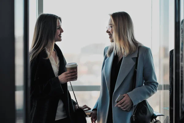 Twee mooie meisjes, een blonde en een brunette, dragen een jas in een glazen lift met een raam. Winkelcentrum of kantoor in de stad. — Stockfoto