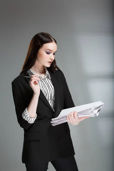 Joven modelo de moda femenina hermosa en un traje de negocios con estilo en un fondo rerom. Sostiene una pila de papeles y una pluma . — Foto de Stock