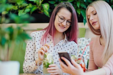 İki genç güzel kız, pembe elbiseli sarışın ve boyalı saçlı esmer, bir kafe masasında oturup, cam kavanoz limonata içmek ve bir akıllı telefon selfie çekmek.