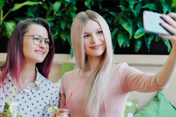 两个年轻的漂亮女孩，一个穿着粉红色连衣裙的金发女郎和一个染发的黑发女郎，坐在咖啡馆的桌子旁，喝着玻璃罐里的柠檬水，在智能手机上自拍. — 图库照片