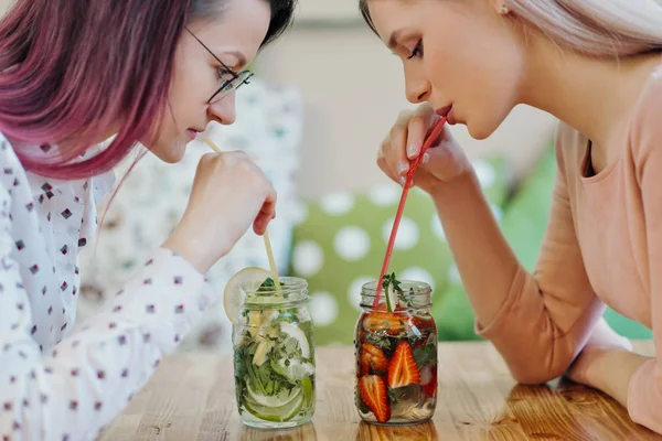 Deux jeunes belles filles, une blonde en robe rose et une brune aux cheveux teints, s'assoient à une table de café, boivent de la limonade dans des pots de verre et prennent un selfie sur un smartphone . — Photo