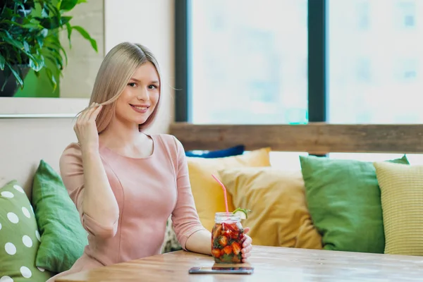 Jeune belle fille blonde dans une robe rose s'assoit à une table de café, boit de la limonade dans un bocal en verre . — Photo