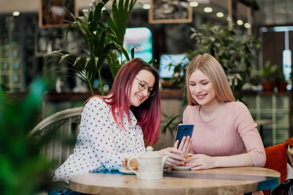 Two young beautiful girls sit in a green cafe at a table. Drink tea with croissants, chatting, laughing and taking pictures on a smartphone.
