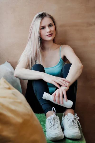 Młoda piękna kobieta atletyczna Blondynka w odzieży sportowej siedzącej przez okno kawiarni na tle drewnianej ściany. — Zdjęcie stockowe