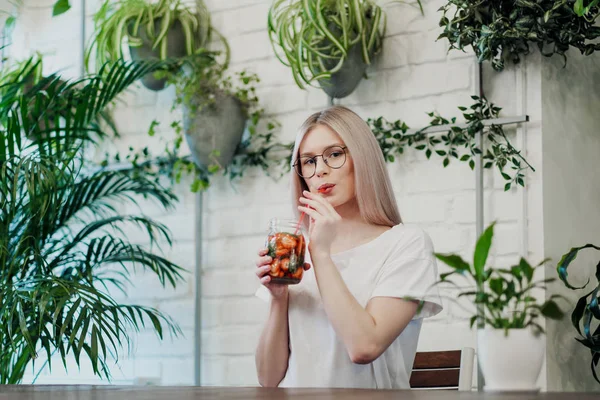 Mladá krásná atletická žena blondýnka v brýlích sedí u stolu v zelené kavárně a pije jahodovou limonádu ze skleněné sklenice. — Stock fotografie