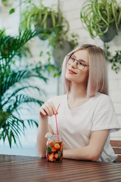 Jeune belle femme sportive blonde à lunettes s'assoit à une table dans un café vert et boit de la limonade de fraises dans un bocal en verre . — Photo