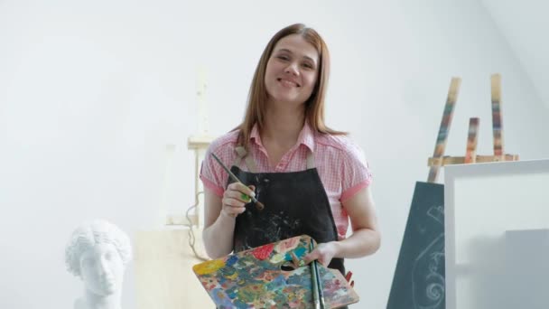 Young beautiful woman painter among easels and canvases in a bright studio. — Stock Video