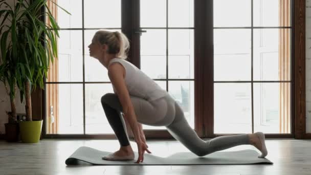 Giovane bella donna atletica bionda in abbigliamento sportivo bianco che fa yoga e si estende su un tappeto grigio sullo sfondo di una grande finestra di edificio a più piani . — Video Stock