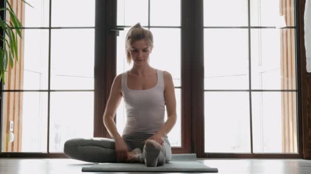 Joven hermosa atlética rubia en ropa deportiva blanca haciendo yoga y estirándose sobre una alfombra gris en el fondo de una gran ventana de edificio de varios pisos . — Vídeos de Stock