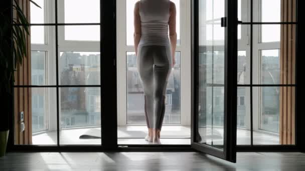 Jovem bela mulher atlética loira em sportswear branco fazendo ioga e alongamento na varanda em casa. Fica nos dedos e executa passos de dança . — Vídeo de Stock