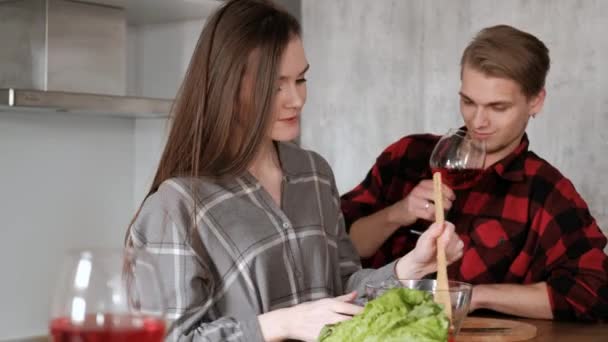 Jovem casal bonito em camisas xadrez estão cozinhando em casa na cozinha. Uma mulher e um homem cortam legumes e fazem uma salada de pimenta, tomate, rabanete em um prato de vidro transparente. . — Vídeo de Stock