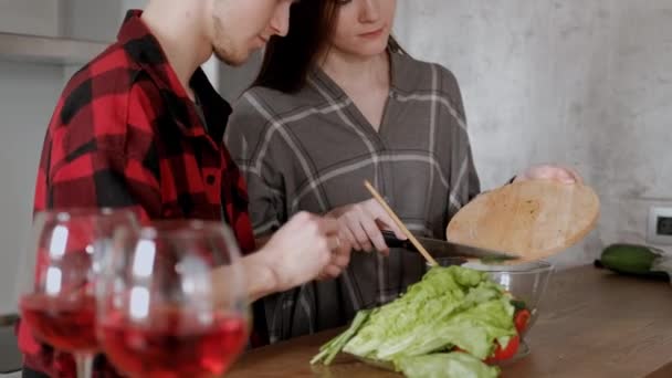 Mladý krásný pár v plovací košili vaří doma v kuchyni. Žena a muž nakrájejte zeleninu a dělají salát z papriky, rajčete, ředkvičky v průhledné skleněné nádobě. — Stock video
